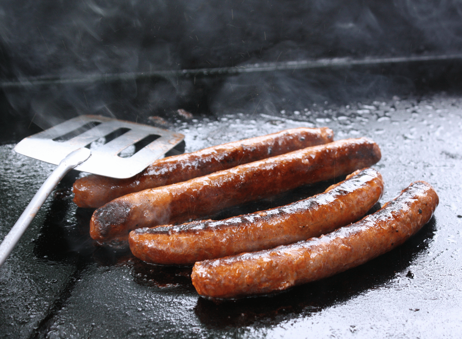 Hotdogs grilling a griddle with a metal spatula