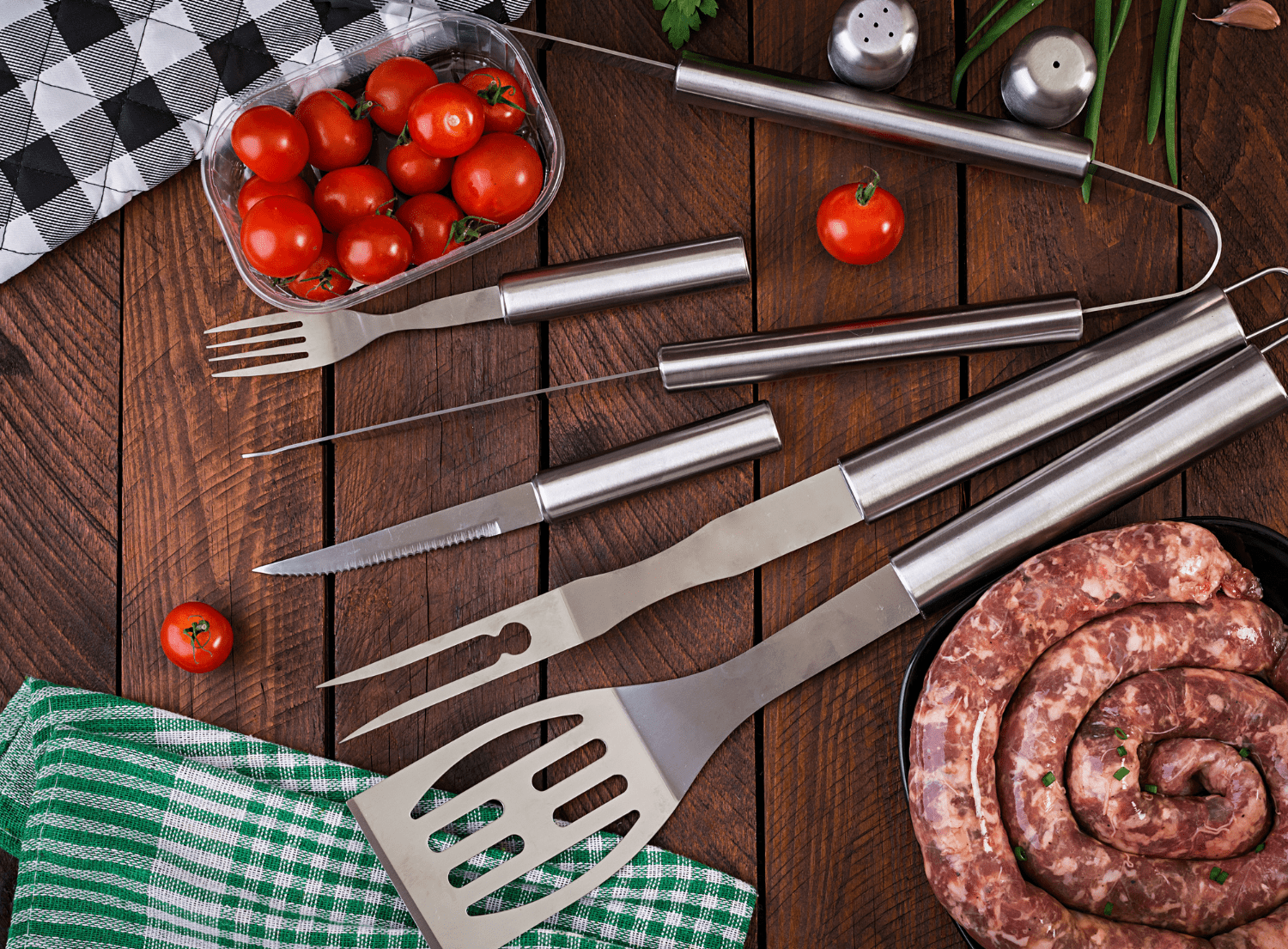 grilling tools, tomatoes and meat laid out on a wooden table